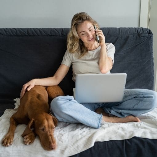 Woman sitting on a couch with a laptop and phone, accompanied by a dog.